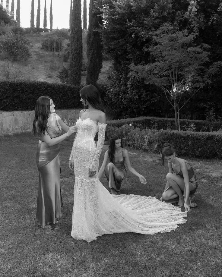 Four women in elegant dresses outdoors on a lawn, one in a wedding gown, two adjusting their dresses, surrounded by trees and bushes.