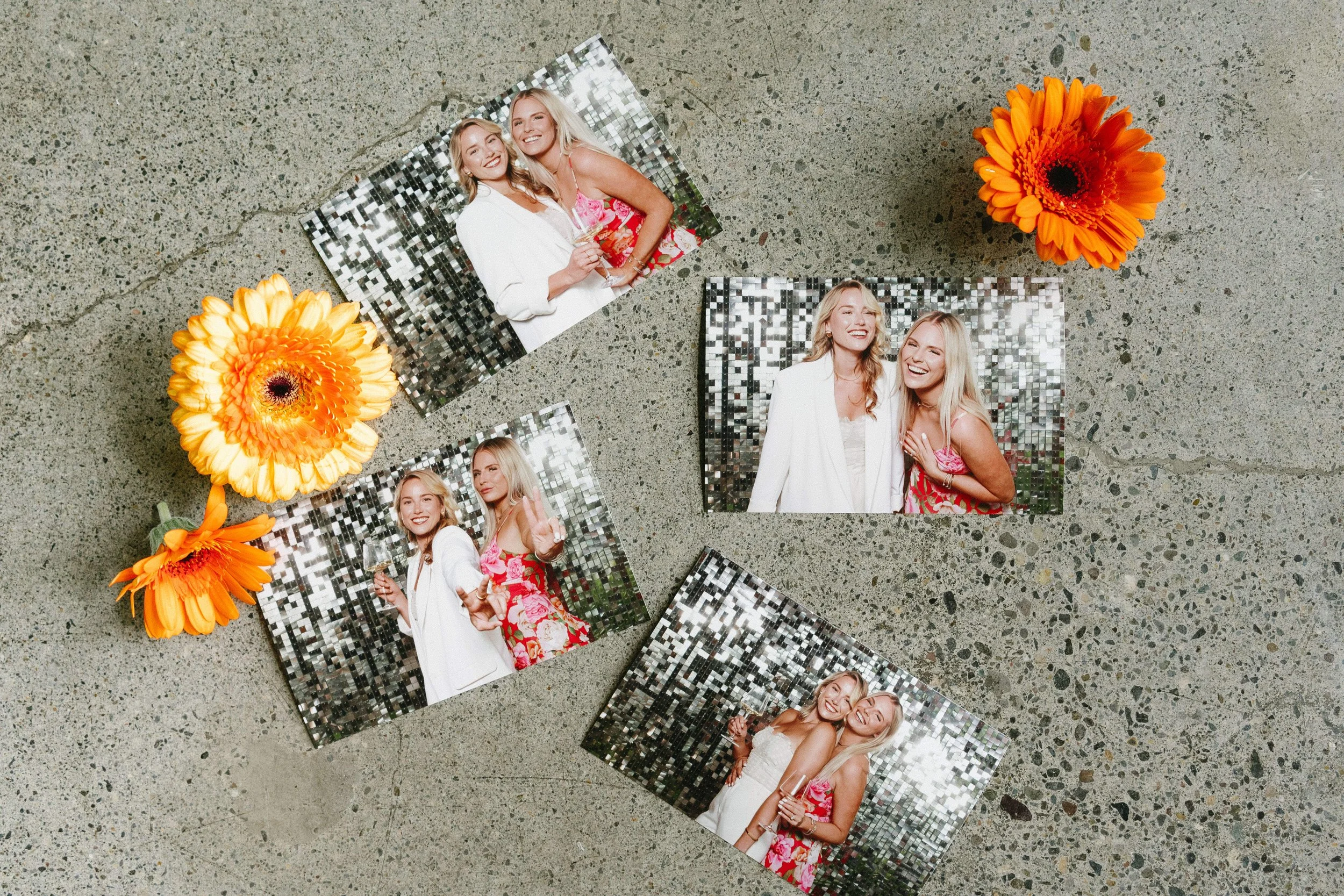 Four printed photographs of two smiling women, one with blonde hair in a white blazer and the other in a pink floral dress, are placed on a gray speckled surface. Two orange gerbera daisies are also on the surface.