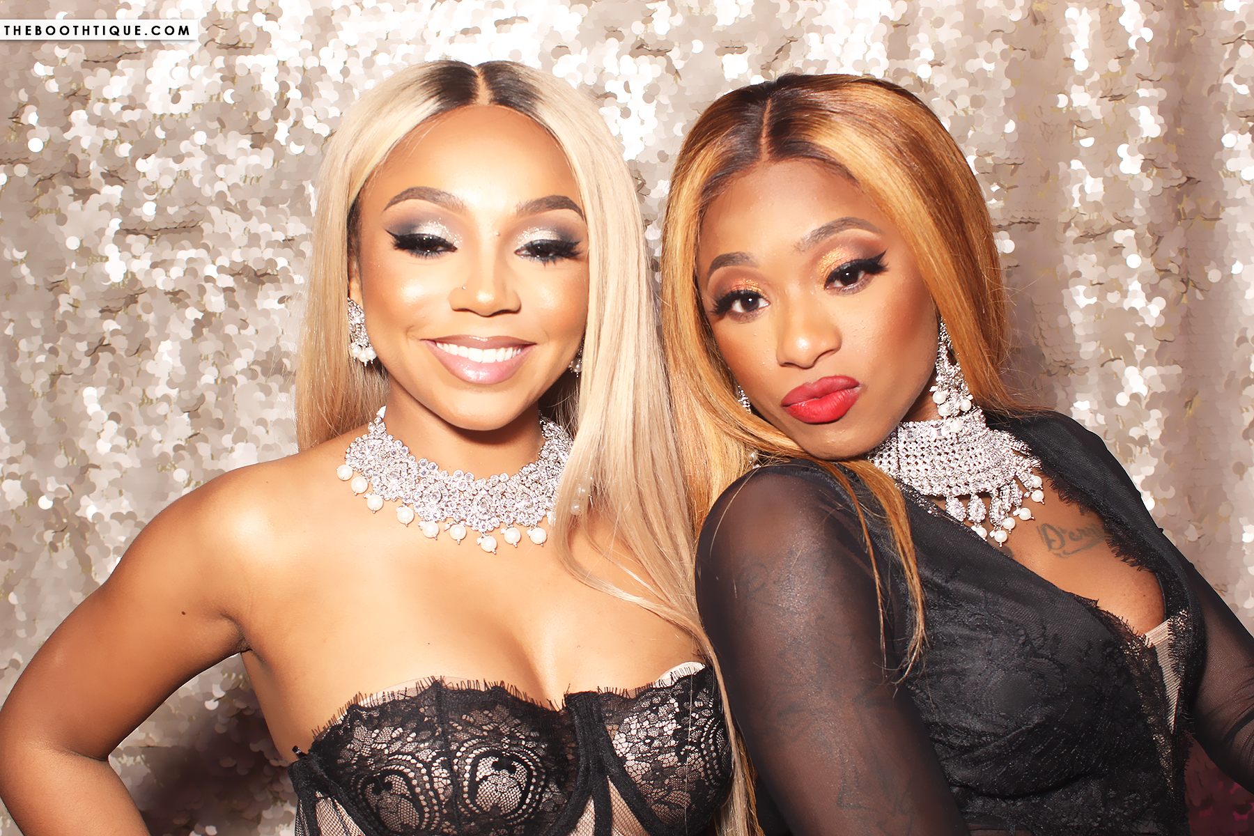 Two women dressed in black lace dresses with jewelry, posing in front of a shiny, sequined backdrop.