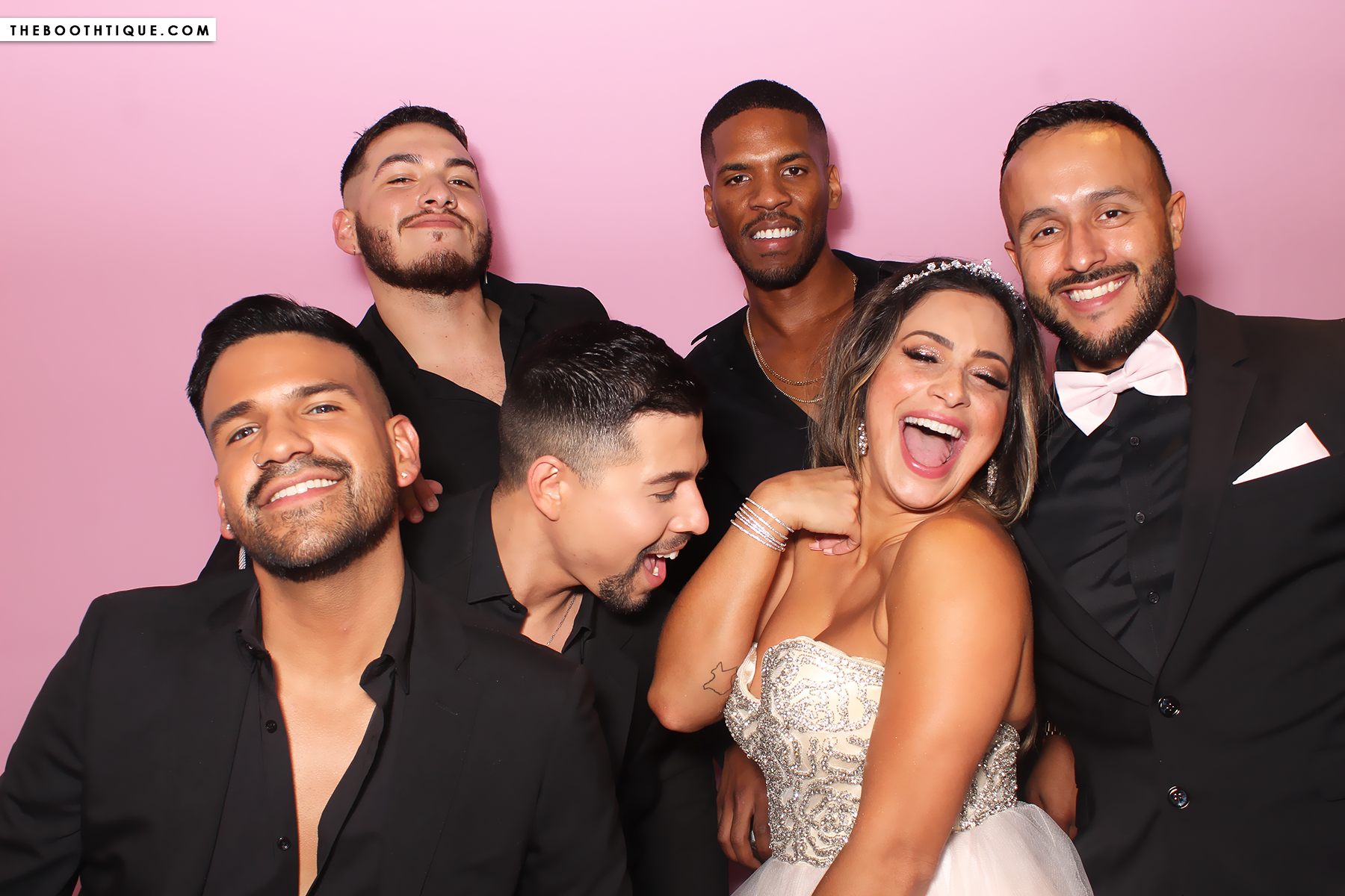 Group of seven friends celebrating in front of a pink background, with one woman wearing a sparkly dress and tiara, laughing joyfully, and six men dressed in black suits, some smiling and some making playful faces.