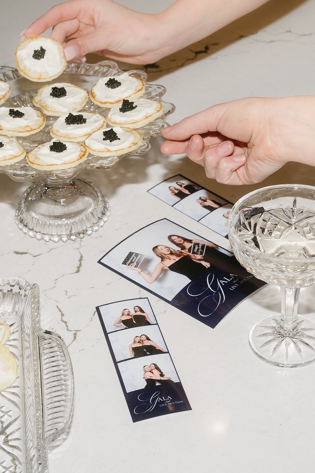 A person is reaching to pick up a small canapé with cream and black caviar, placed on a glass cake stand with other similar appetizers. Various event photographs and promotional brochures for a gala are laid out on a marble table, along with a glass 