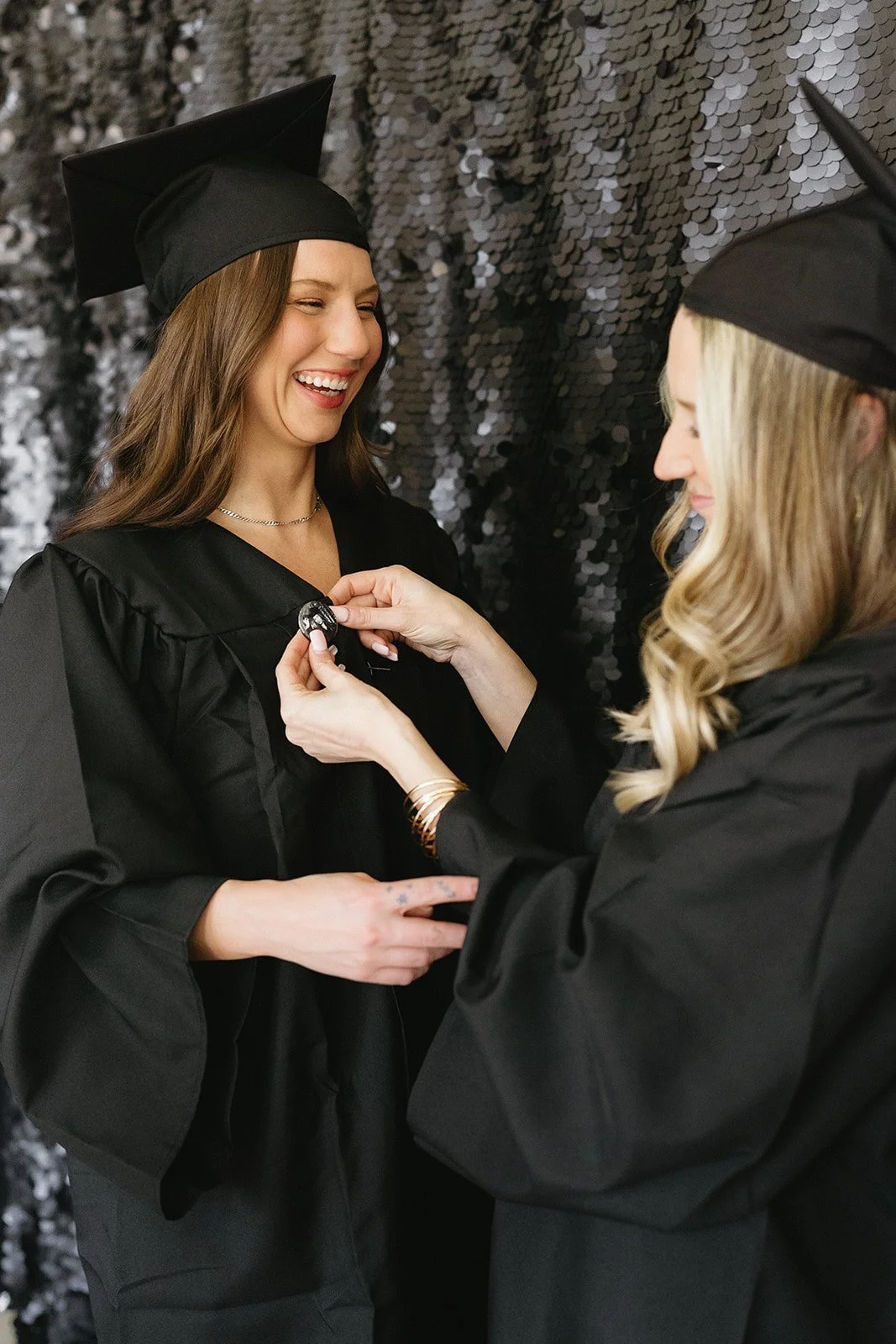 Two women in black graduation gowns and caps, one is pinning a medal or badge on the other's gown during a graduation ceremony.