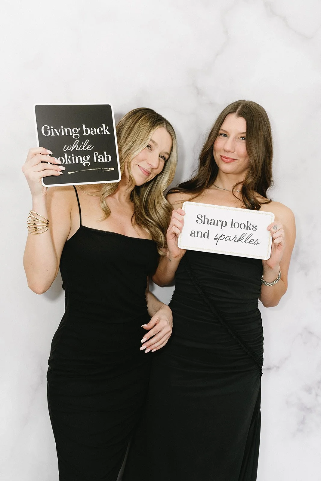 Two women in black dresses holding signs with positive messages about looks and personality, standing against a white marble background.