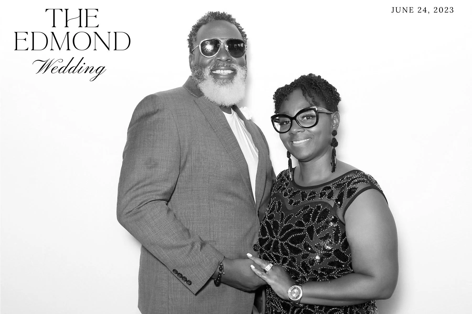 Black-and-white photo of a smiling couple dressed elegantly, holding hands, with the man wearing dark sunglasses and the woman wearing large glasses and earrings, standing against a plain white background. The text in the top left corner reads 'The E