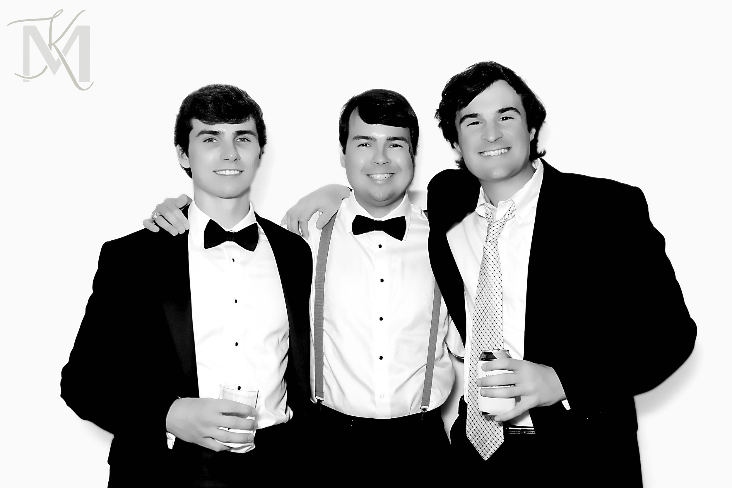 Three young men dressed in formal tuxedos and bow ties, smiling, with drinks in their hands, standing together against a plain background.