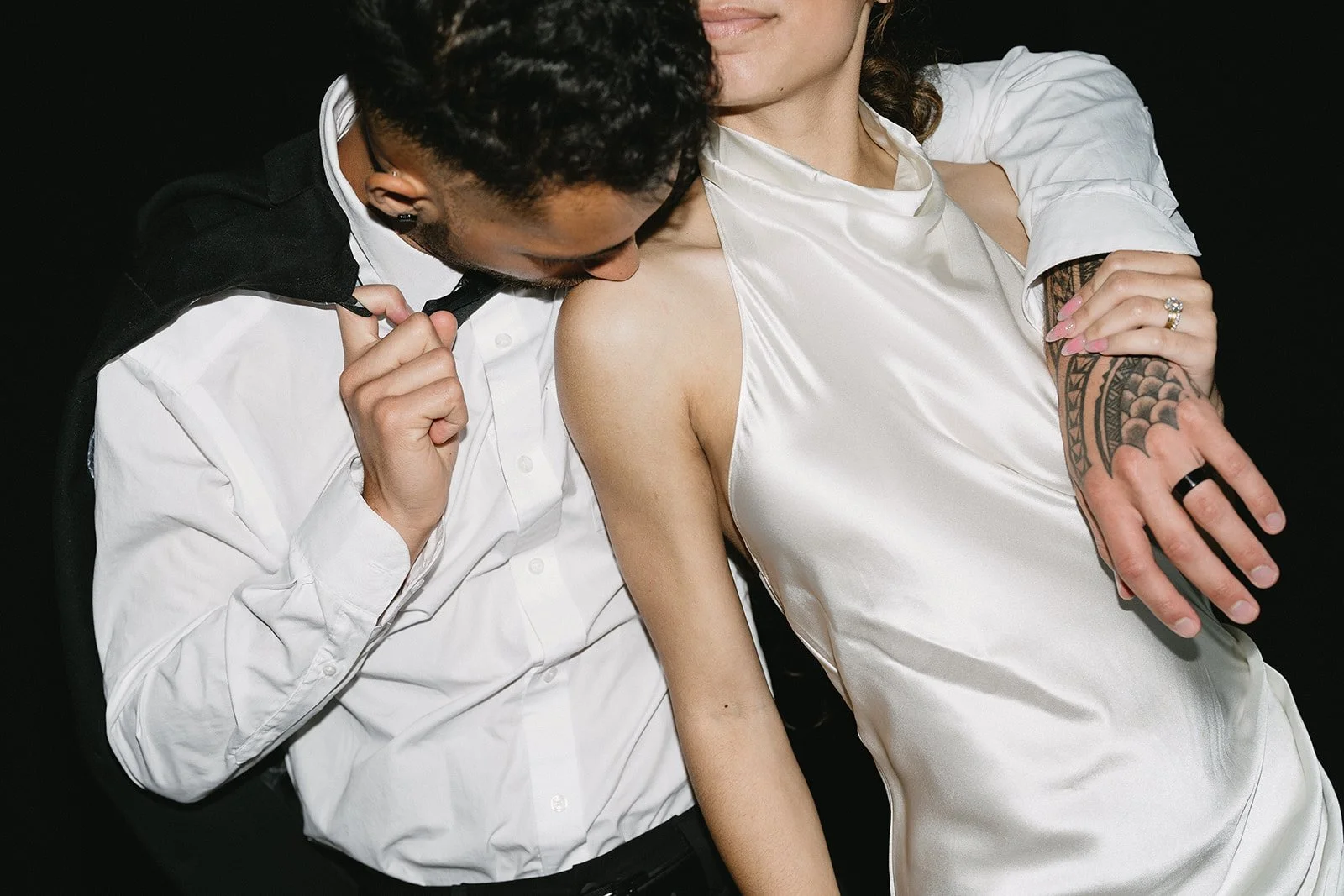 A man in a white shirt and black jacket leans in to kiss a woman's shoulder, who is wearing a satin white dress and has a tattoo on her arm. The woman's hand shows a ring and pink nails.