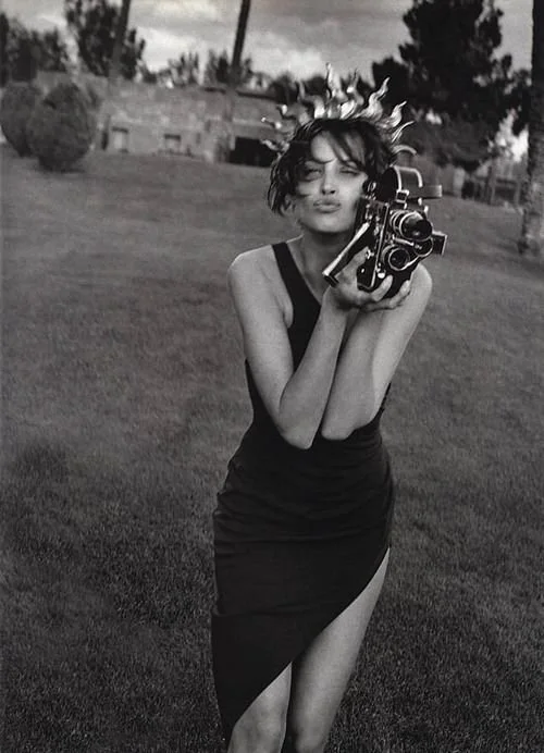 A woman in a black dress holding a vintage camera, standing on a grassy field with trees and a building in the background, black-and-white photo.