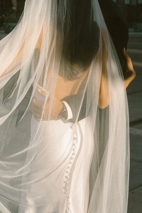 A bride in a white wedding dress with buttons, partially covered by a sheer veil, standing outdoors.