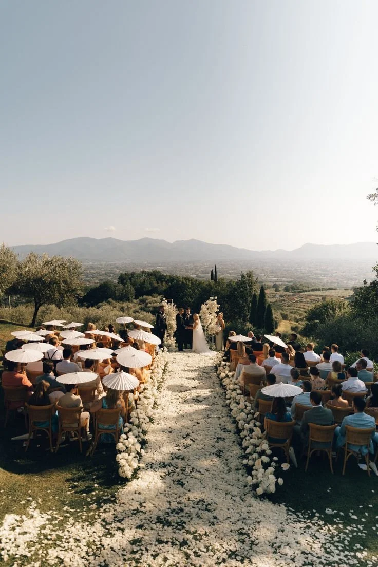 Outdoor wedding ceremony with guests seated under umbrellas, officiant and bride and groom at the altar, surrounded by flowers on a scenic hillside.