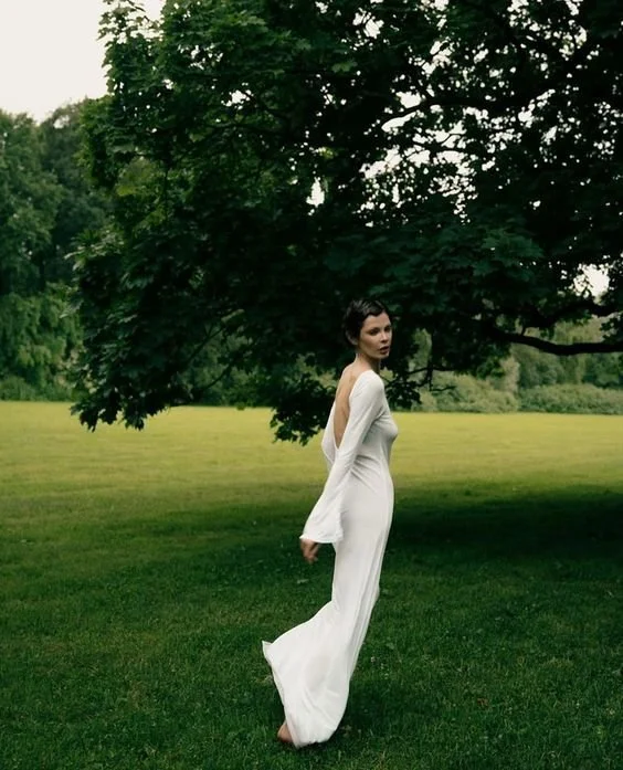 A woman in a white dress standing on grass in a park with a large leafy tree overhead.