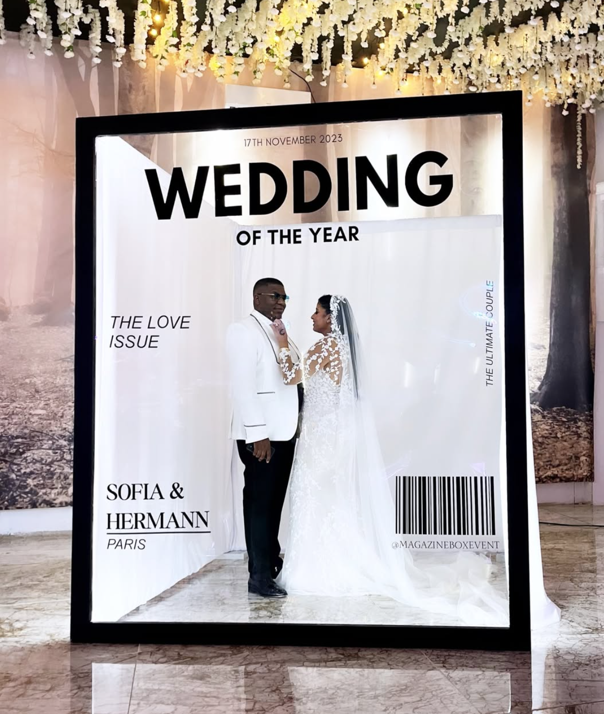 A framed photo of a bride and groom inside a decorative display at a wedding event, with floral ceiling decorations and a barcoded label at the bottom right. The event is titled "Wedding of the Year" held on November 17, 2023, featuring married couple Sofia and Hermann from Paris.