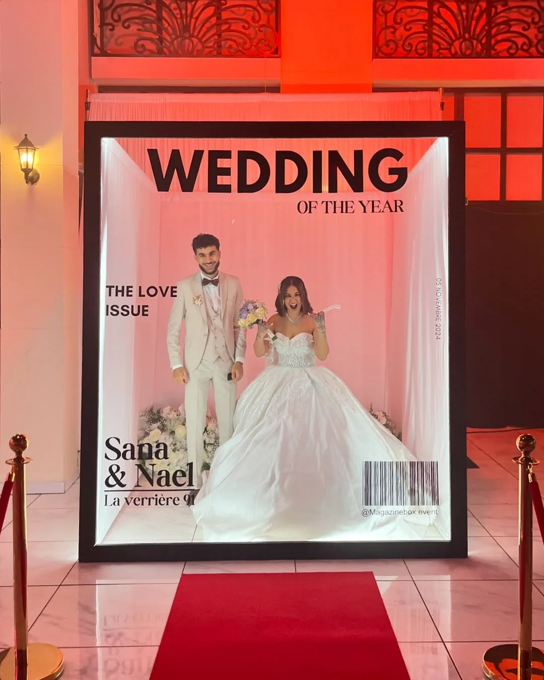A large illuminated frame displays a poster for a wedding of the year, featuring a smiling bride and groom in wedding attire. The groom is in a white suit with a bow tie, and the bride in a white wedding gown holding a bouquet of flowers. The poster includes text for the bride and groom's names, Sana and Nael, and mentions a love issue, with the date November 2024. The display is set indoors with warm lighting and a red carpet in front.