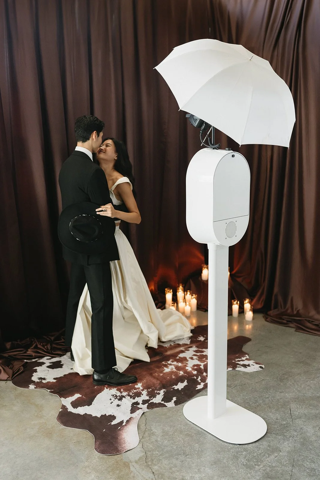 A couple dressed in wedding attire embracing and smiling at each other on a decorated backdrop with candles in the background during a photo booth session.