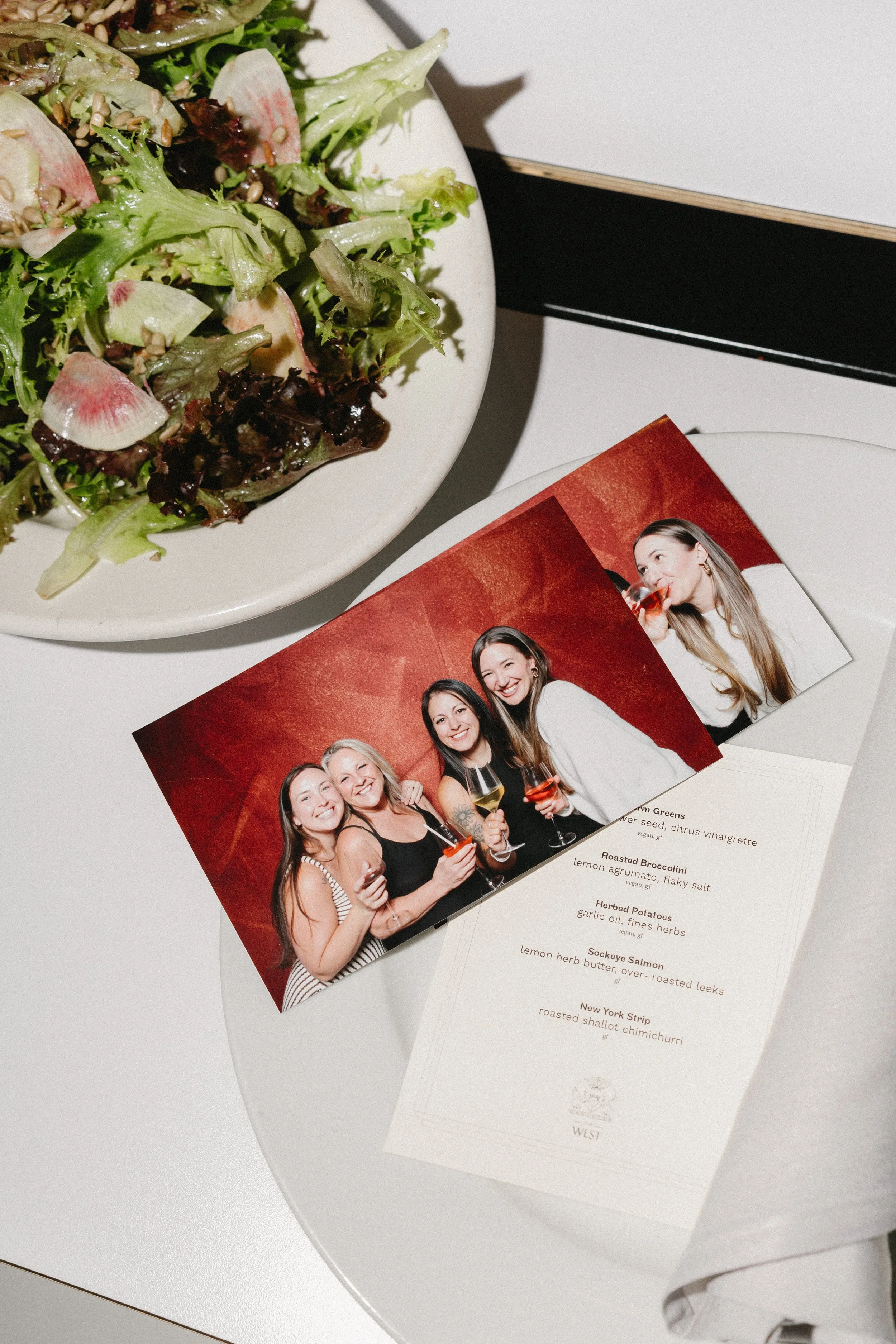 A plate of mixed green salad with radish slices, a photo of four women smiling and holding drinks, and a menu with dishes including roasted broccolini, herbed potatoes, sockeye salmon, and New York strip steak.