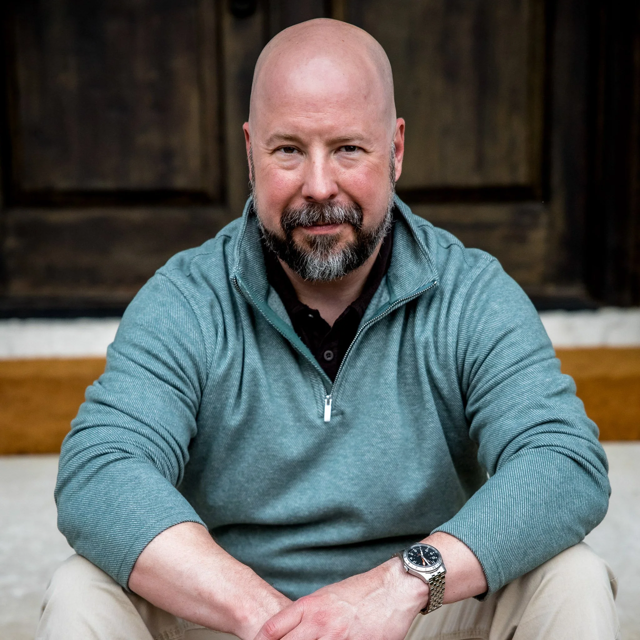 A middle-aged man with a beard, wearing a teal quarter-zip sweater and a wristwatch, sitting in front of a wooden background.