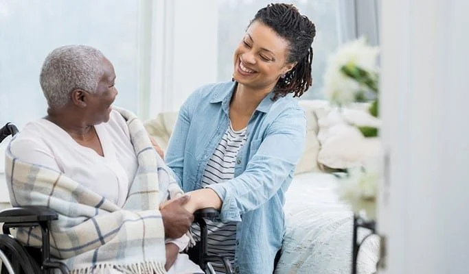 A young woman smiling and holding hands with an elderly woman in a wheelchair in a bright, cheerful room.