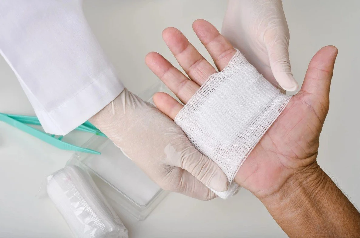 A healthcare worker in gloves bandages a patient's hand with a white gauze pad.