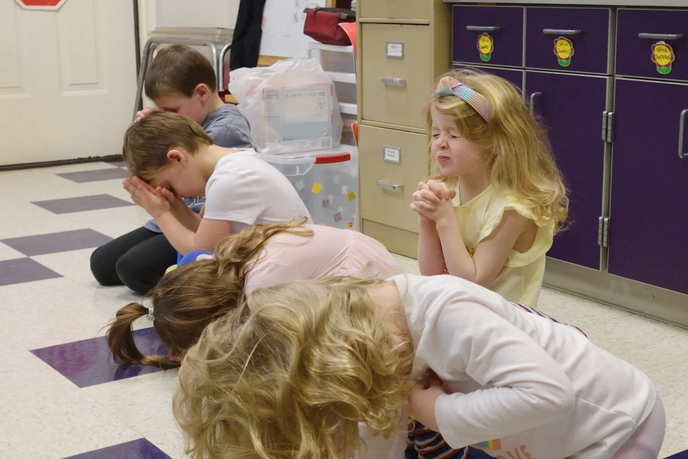 Five children kneeling on the floor in a classroom, with their heads bowed and hands clasped as if praying, a girl with red hair and a rainbow headband looks distressed.