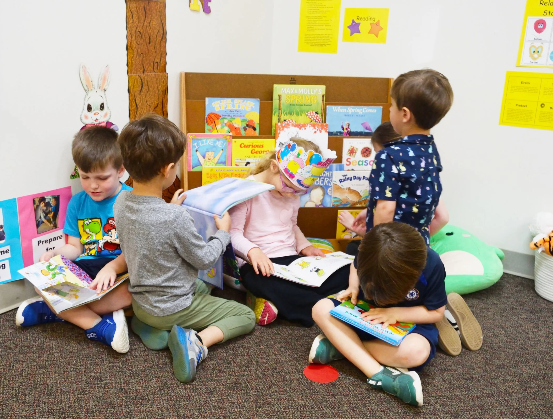 Group of young children reading books together in a classroom corner with colorful posters and plush toys.