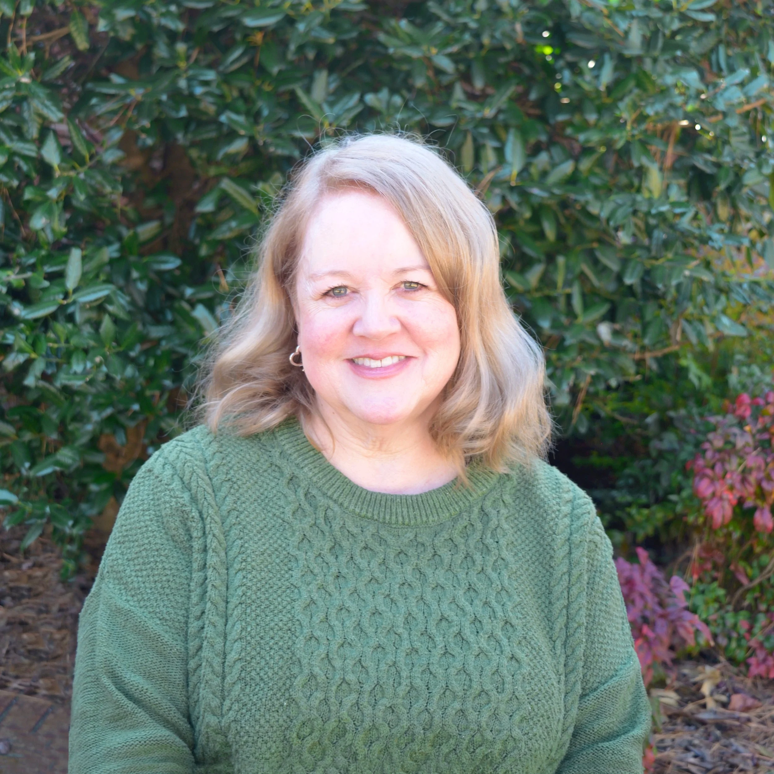 A woman with blonde curly hair smiling outdoors in front of a brick building with an arched doorway.