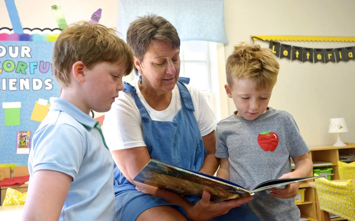 A woman reading a book to two young boys in a classroom with colorful decorations and a bulletin board.