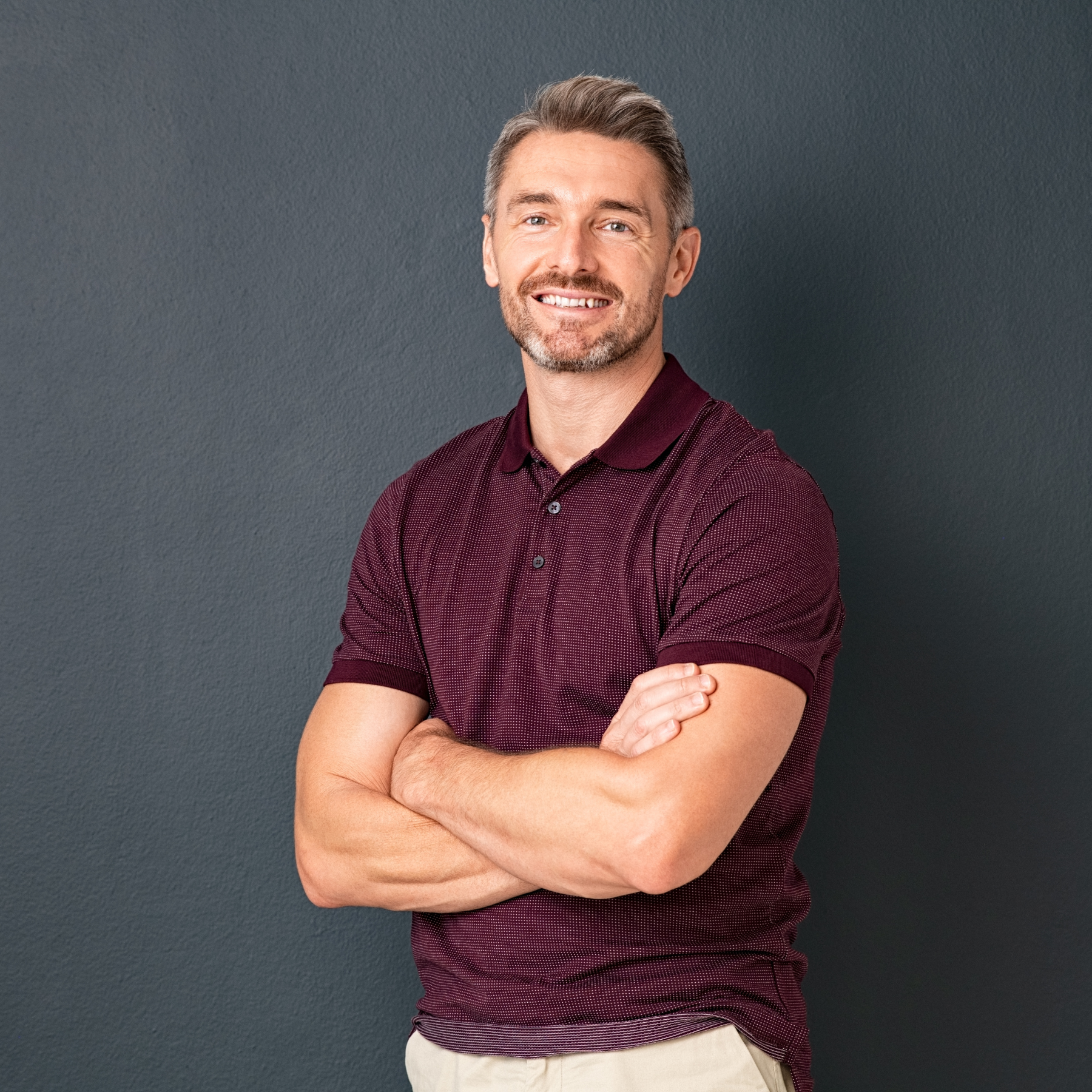A middle-aged man with gray hair and a beard, smiling, standing with arms crossed, wearing a maroon polo shirt, against a dark gray background.