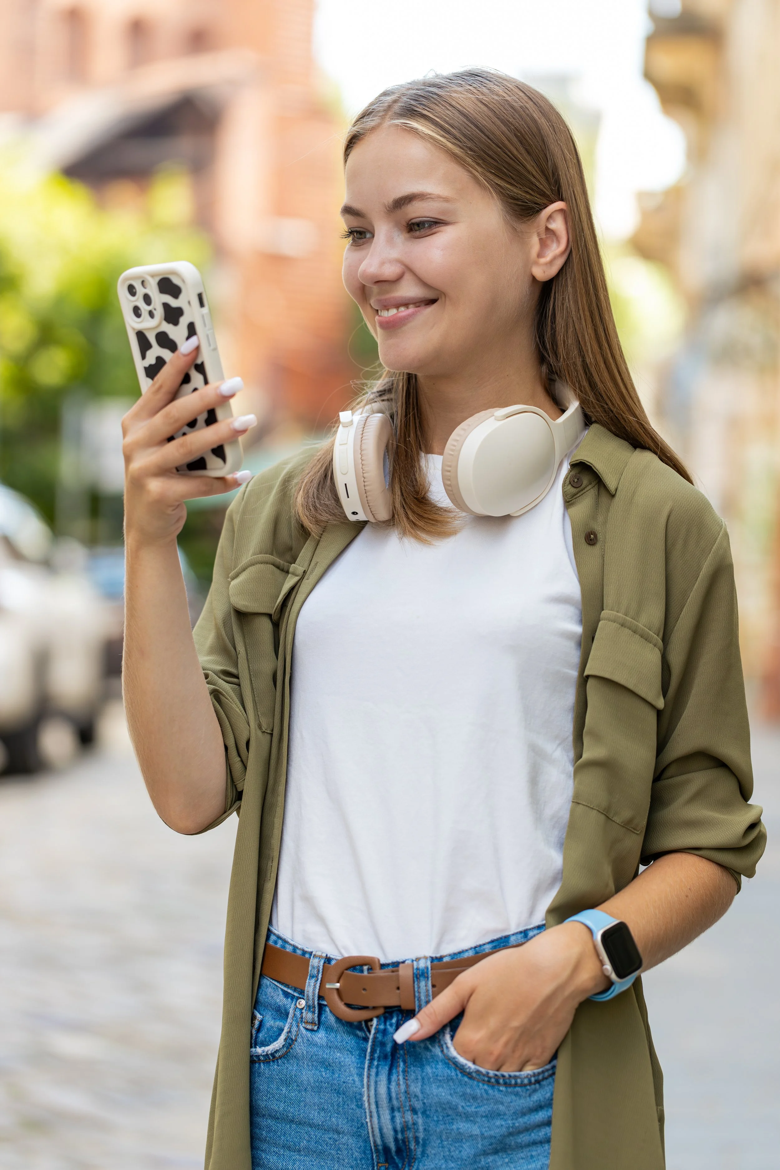 Young woman in her 30s walking through the city with headphones around her neck, smiling as she looks at her phone—capturing the calm, confident energy of using the FocusFlow productivity app on the go.