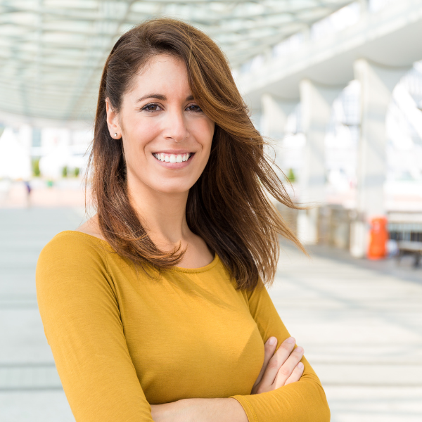 A woman with brown hair, wearing a mustard yellow top, smiling with arms crossed, standing outdoors with a modern architectural backdrop.
