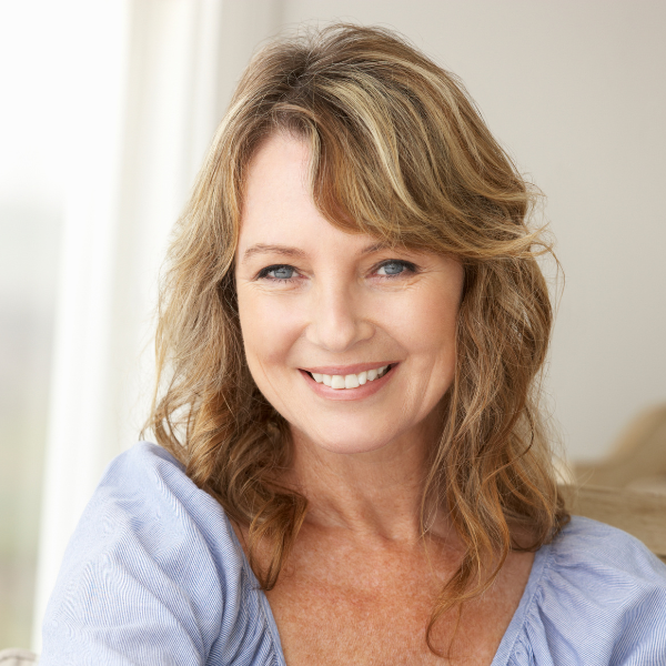A smiling woman with shoulder-length wavy light brown hair and blue eyes, wearing a light blue top, sitting indoors in front of a window with white curtains.