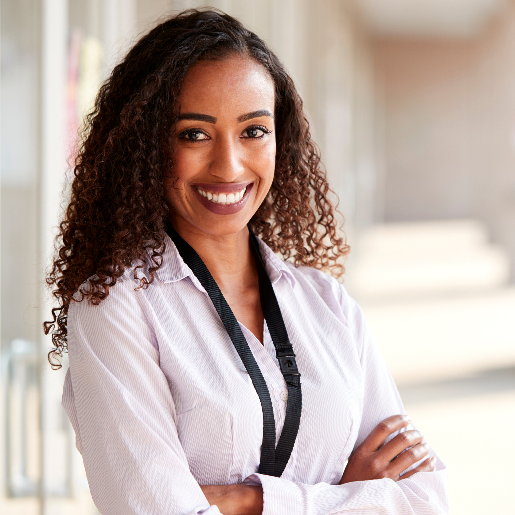 A smiling woman with curly hair and a white shirt standing with arms crossed, wearing a camera strap around her neck in an indoor setting.