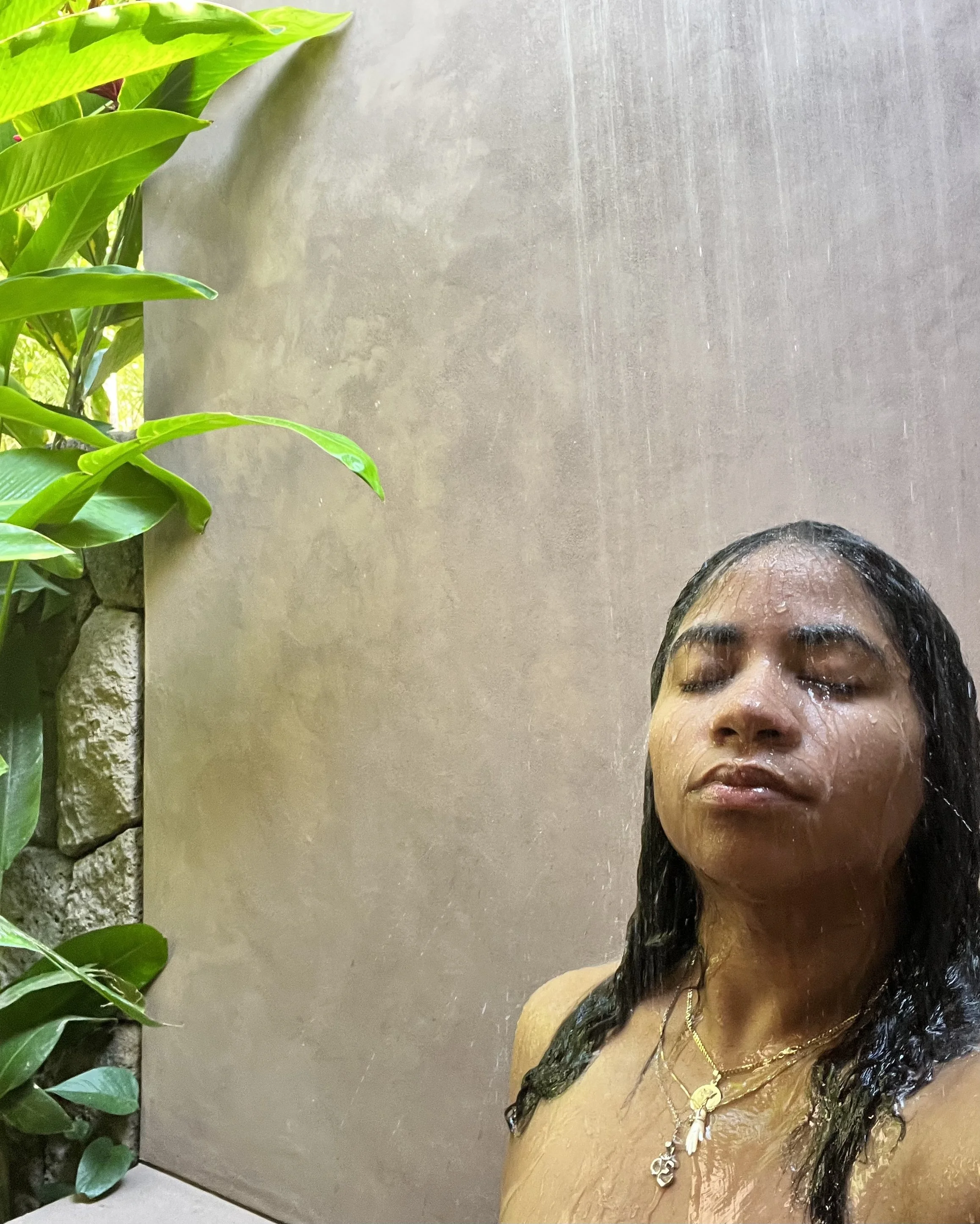 A woman with long dark hair and multiple necklaces is taking a shower, with water water streaming down her face and body. Green leaves of a plant are visible on the left side of the image.
