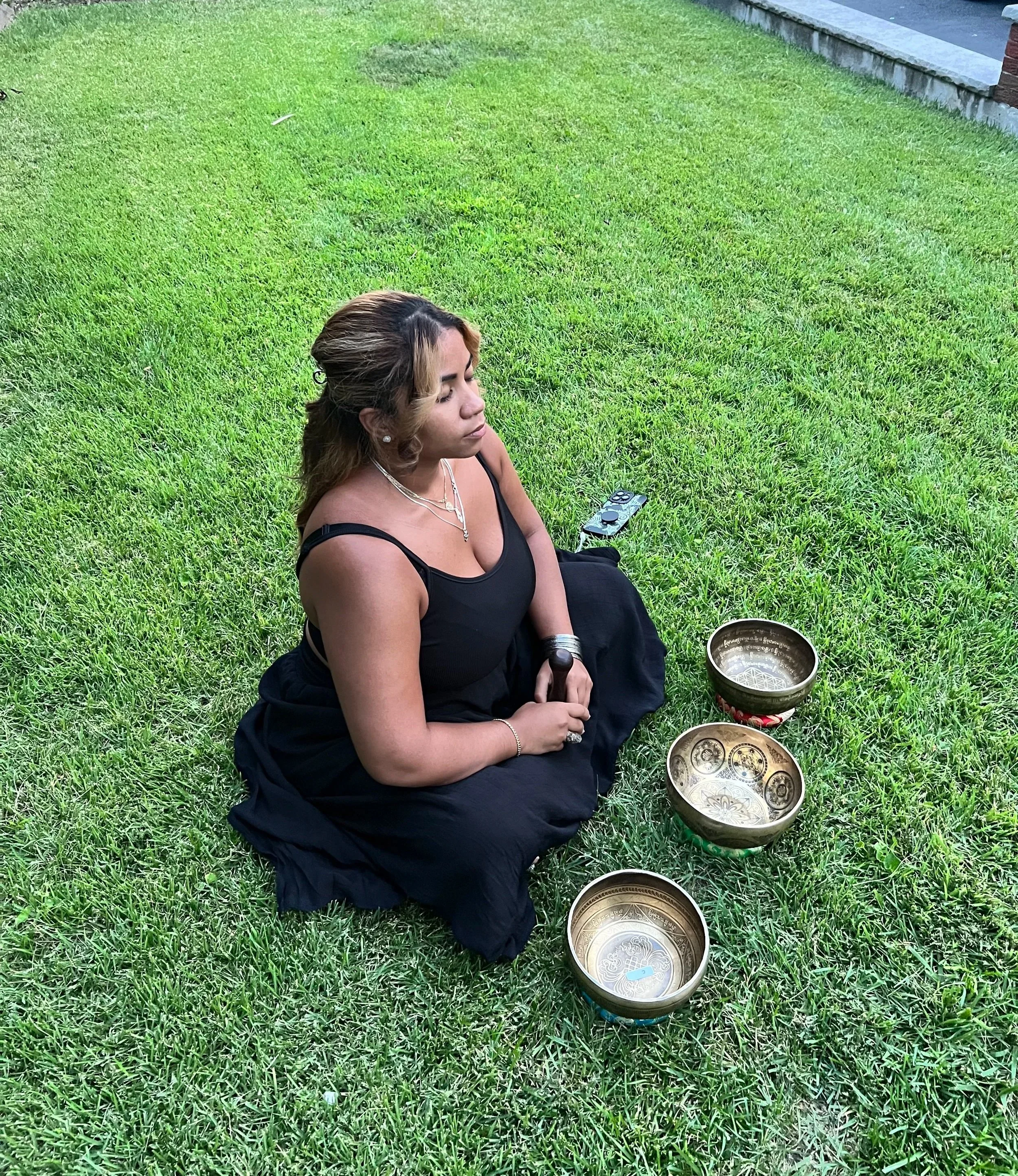 A woman sitting cross-legged on a grassy lawn with three Tibetan singing bowls in front of her, a small remote on the ground nearby, and a brick building with a concrete step in the background.