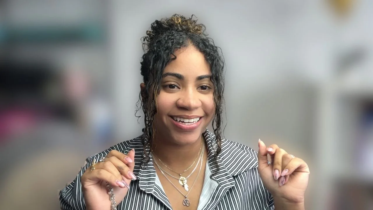 A woman with curly hair smiling and holding her hands near her shoulders, wearing layered necklaces and a striped shirt.