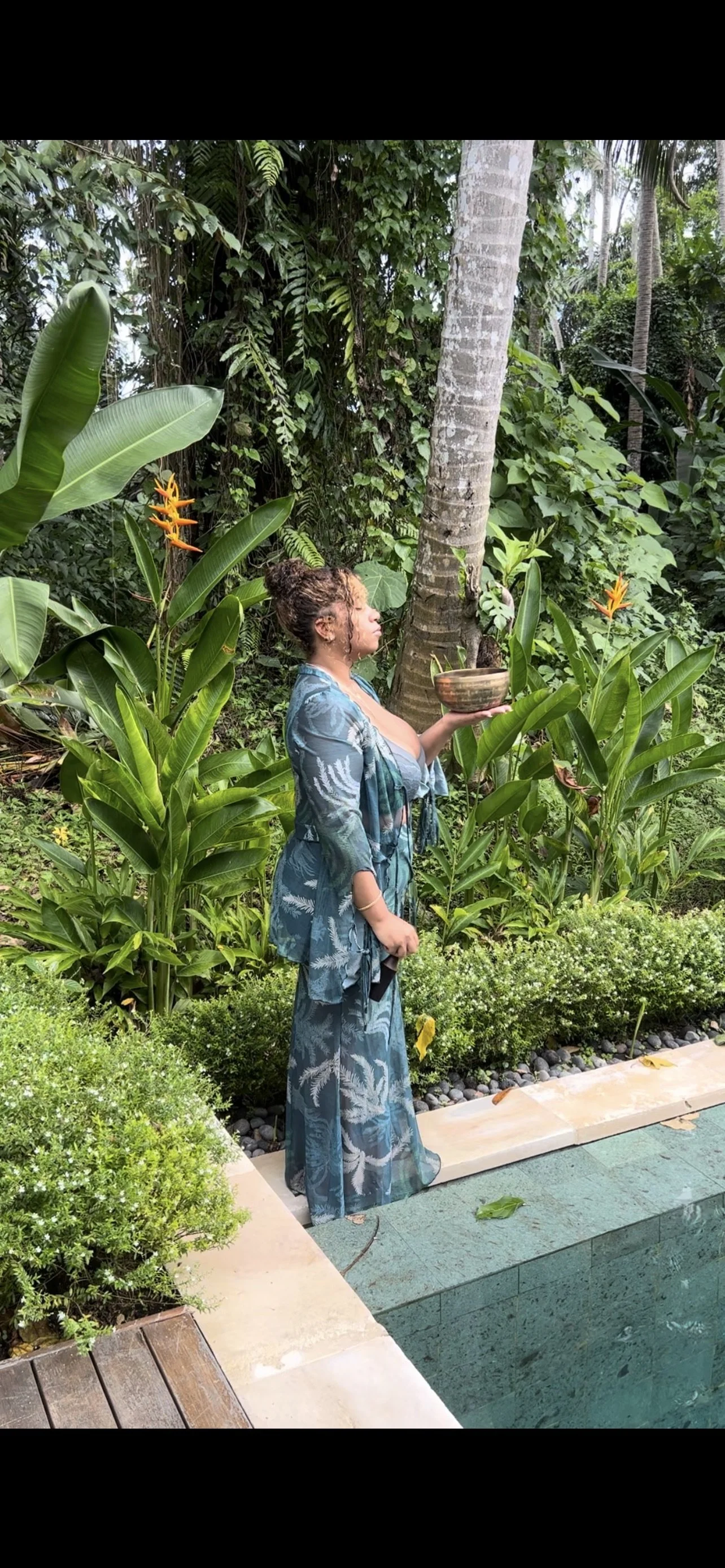 A woman in a teal-patterned dress stands by a pool, holding a bowl, surrounded by tropical plants and trees, with a lush background of greenery.