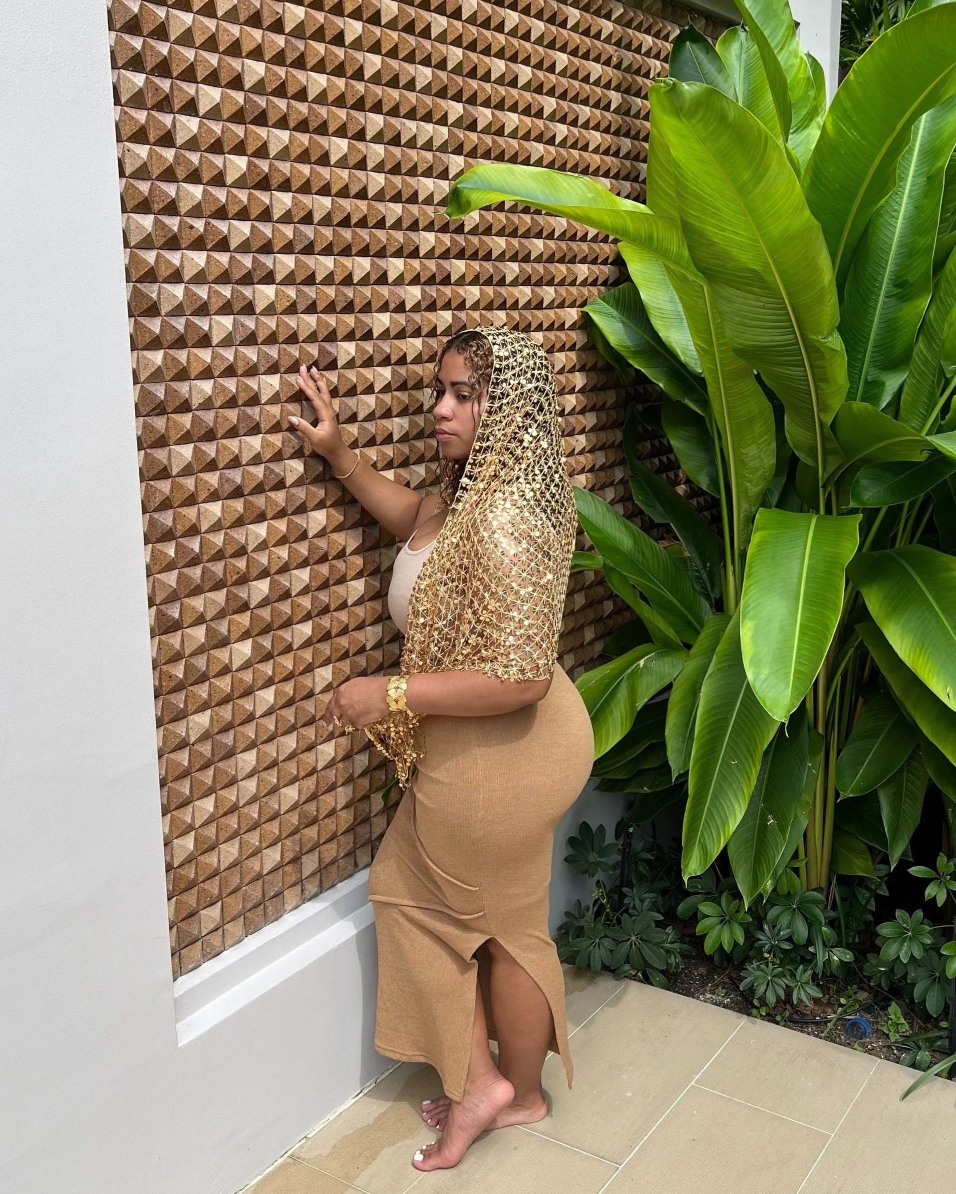 A woman with curly hair standing barefoot next to a textured wooden wall and large green plants, wearing a beige dress and a gold shawl.