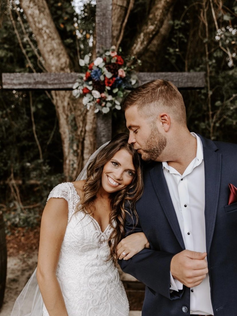 A newlywed couple, with the groom kissing the bride on her forehead, standing in front of a wooden cross decorated with flowers in an outdoor setting.