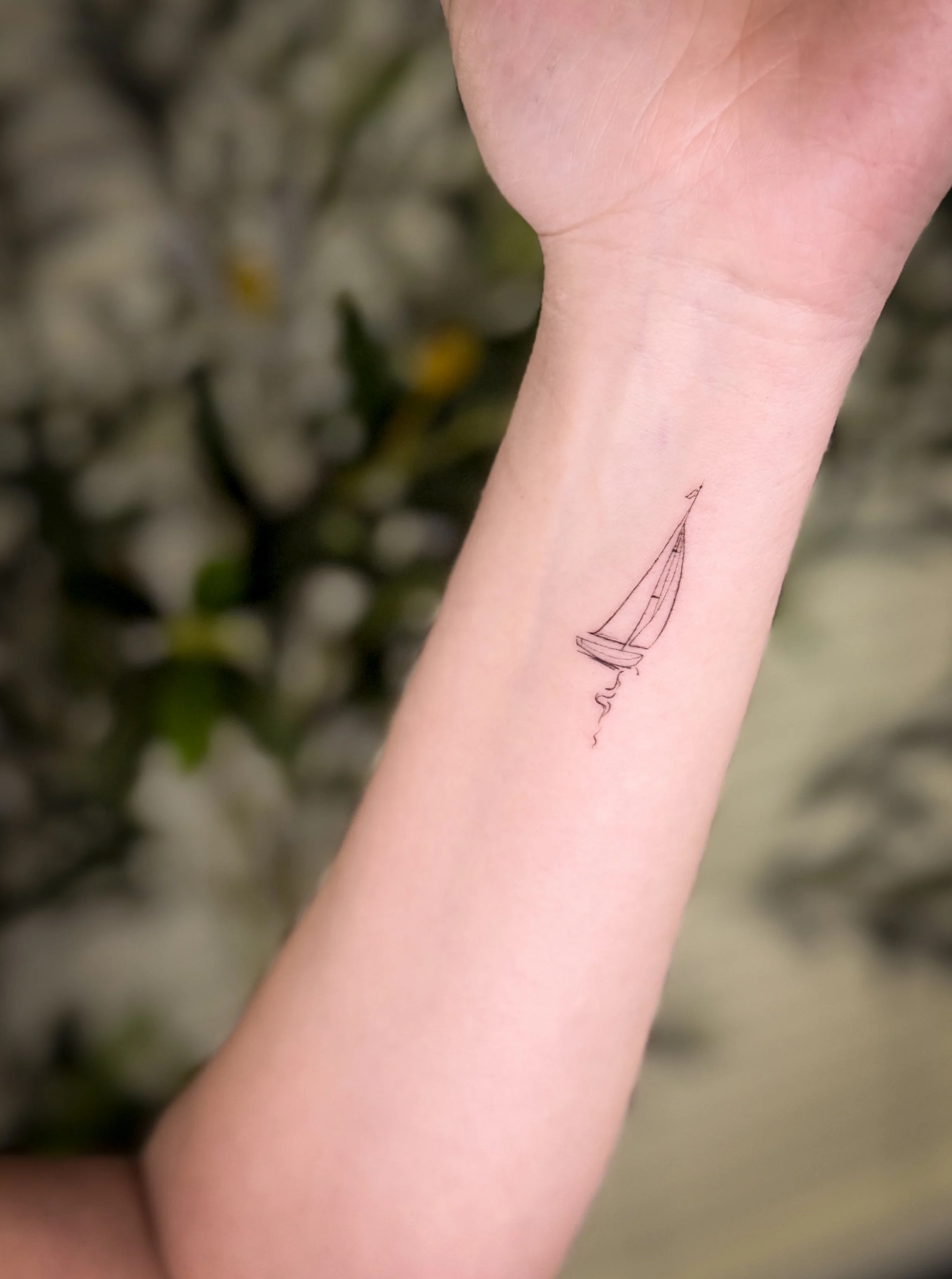 An inner forearm tattoo of a minimalistic sailboat with a face profile, including a sail and a small wave.