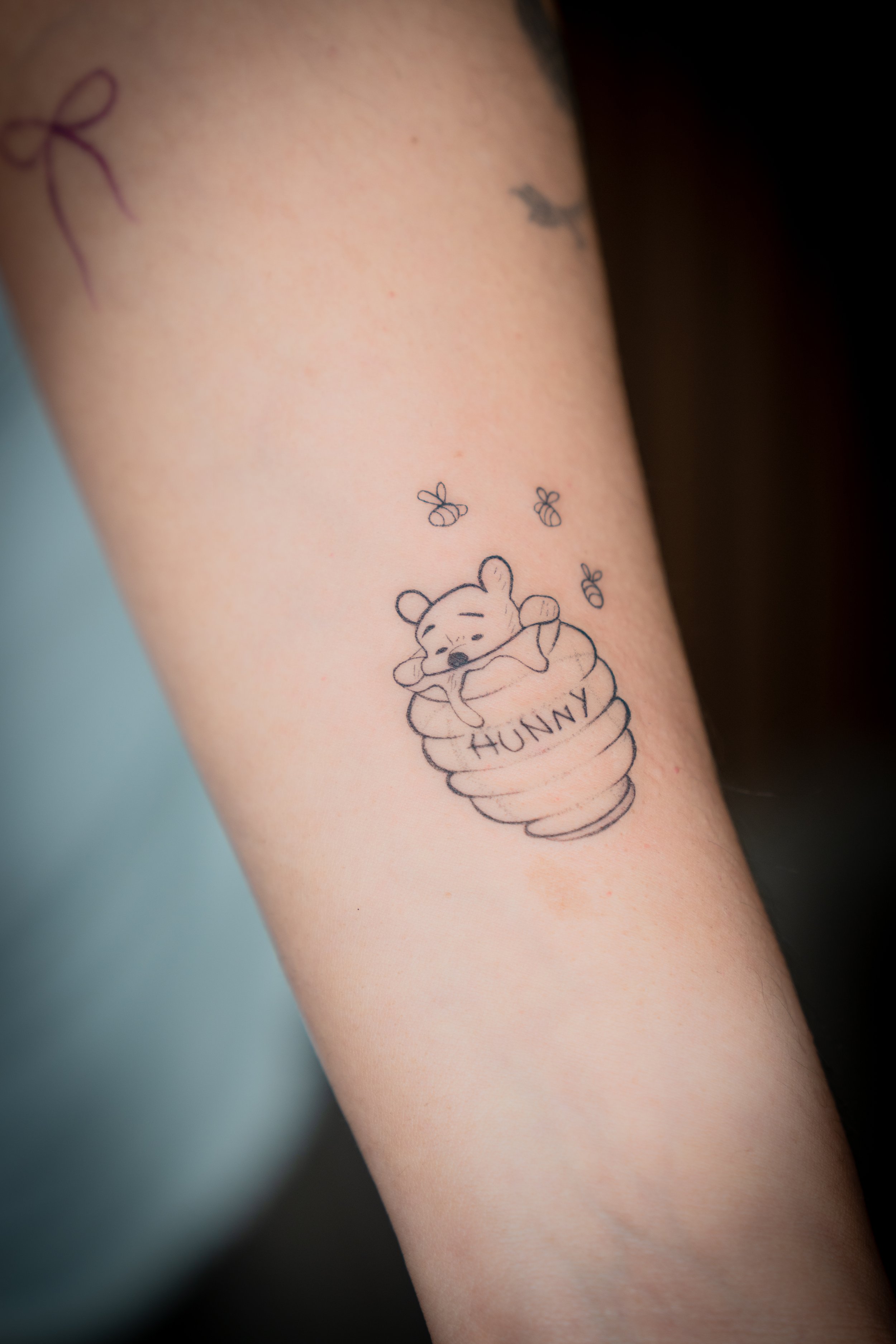 Tattoo of a cute bear resting in a honey pot labeled 'HUNNY,' with small bees flying around.