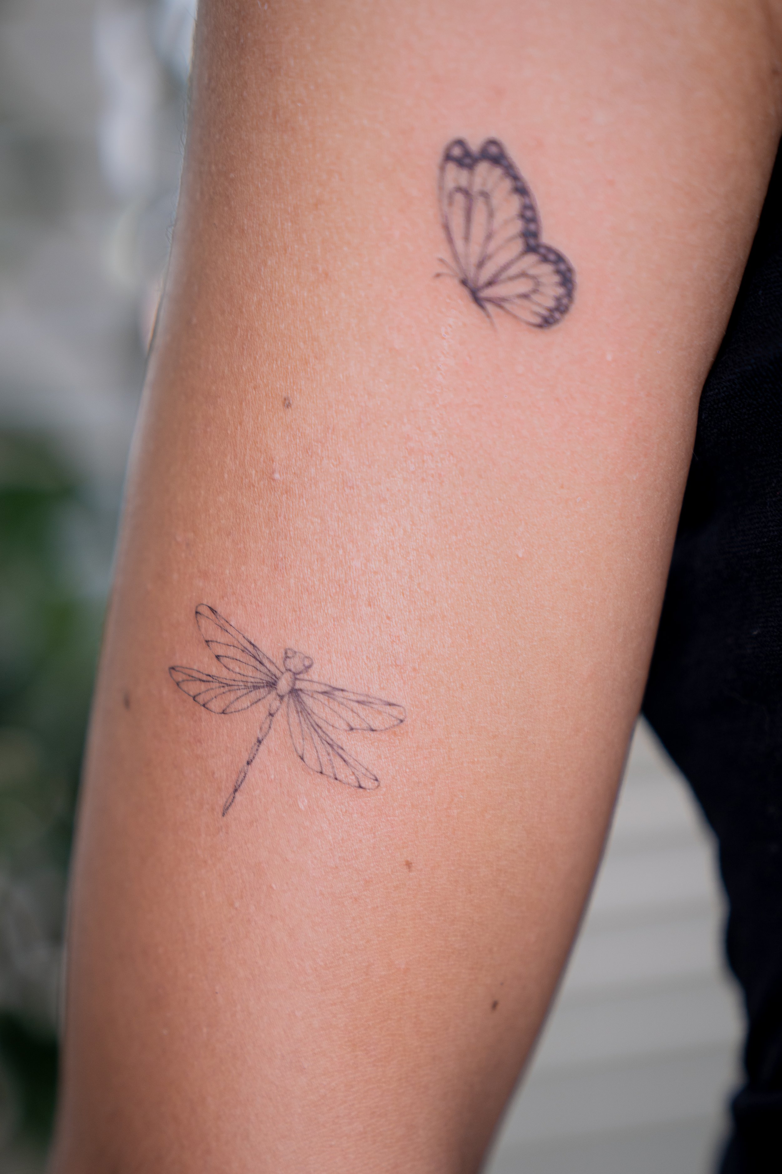 Two delicate line-drawn tattoos: a butterfly on the upper arm and a dragonfly on the forearm.