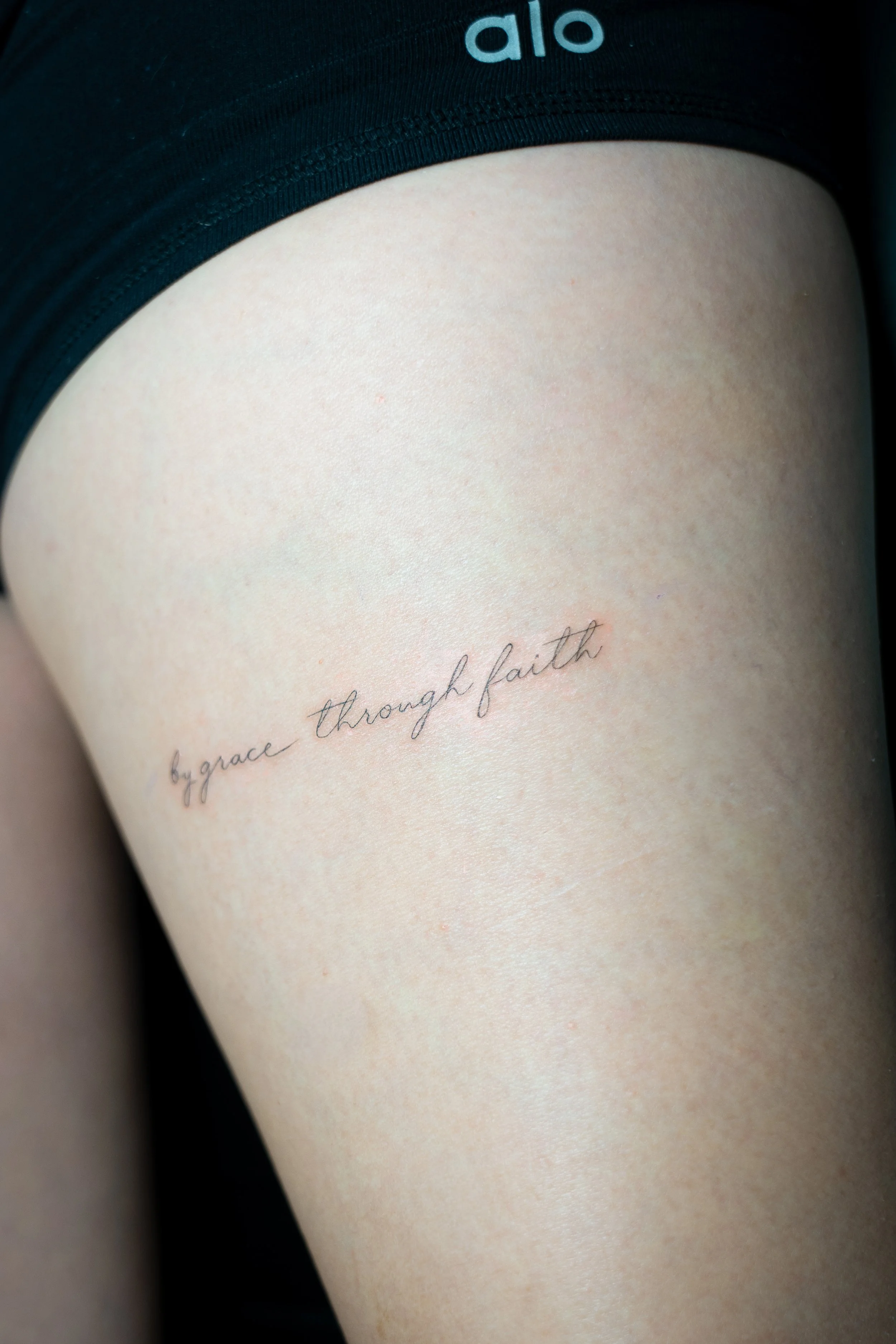 Close-up of a person's upper thigh with a tattoo that reads 'by grace through faith' in cursive script.
