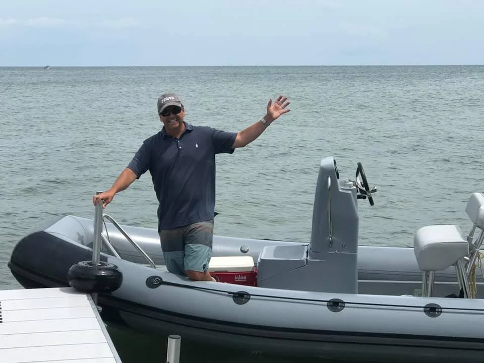 Man standing on a small motorized boat in the water, smiling and waving at the camera.