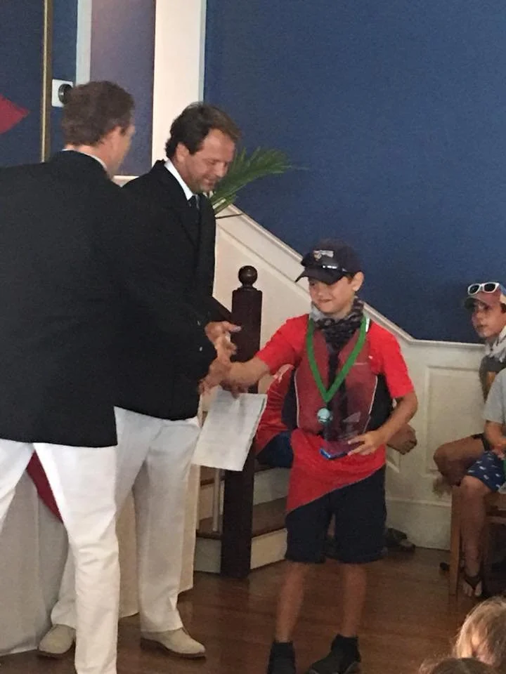 A child wearing a medal, cap, and sunglasses receives an award from two adults in formal attire during an indoor event.