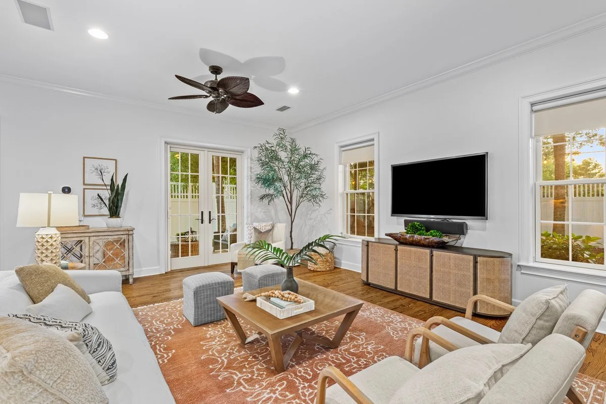 Living room with white walls, hardwood flooring, a large flat-screen TV, a ceiling fan, a sliding glass door, and windows with blinds, decorated with plants and cozy furniture.