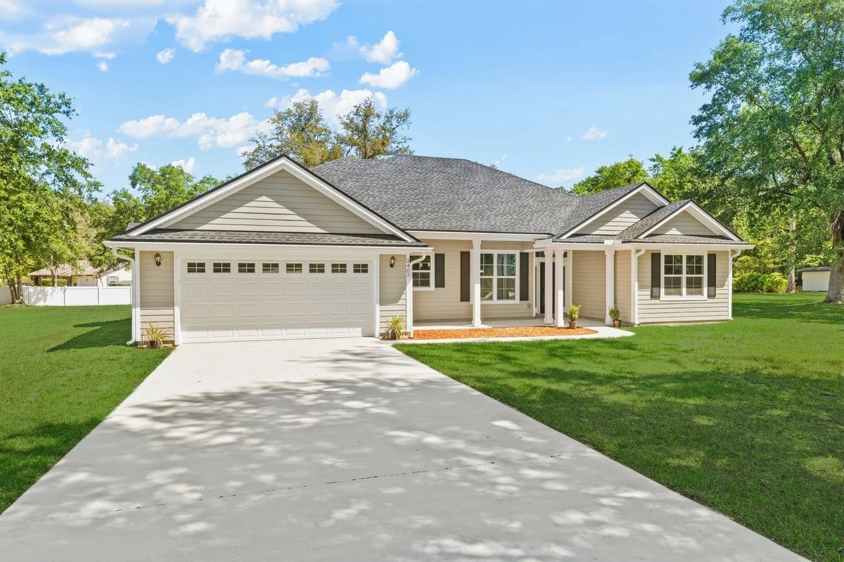 A single-story house with beige siding, dark shutters, and a two-car garage, surrounded by green lawn and trees on a sunny day.
