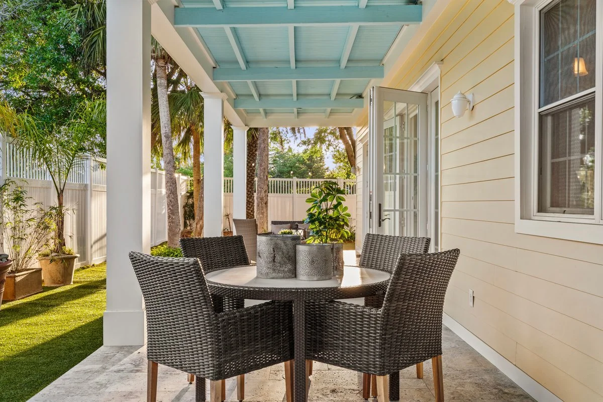 Covered outdoor patio with a round wicker table and six chairs, potted plants, and a glass door leading inside a house with yellow siding, white trim, and a wall-mounted light fixture. Green trees and a white fence are visible in the background.