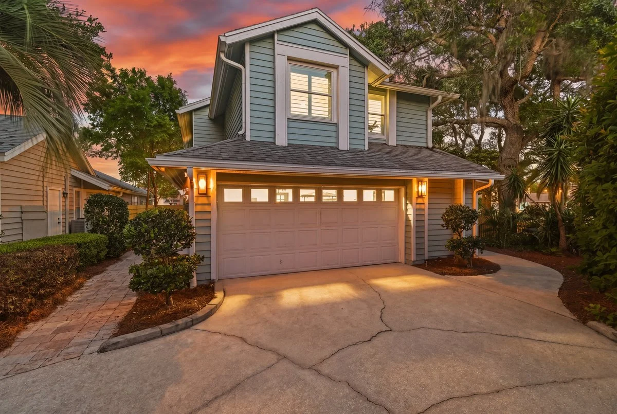 A two-story house with a garage door, surrounded by trees and bushes, during sunset.
