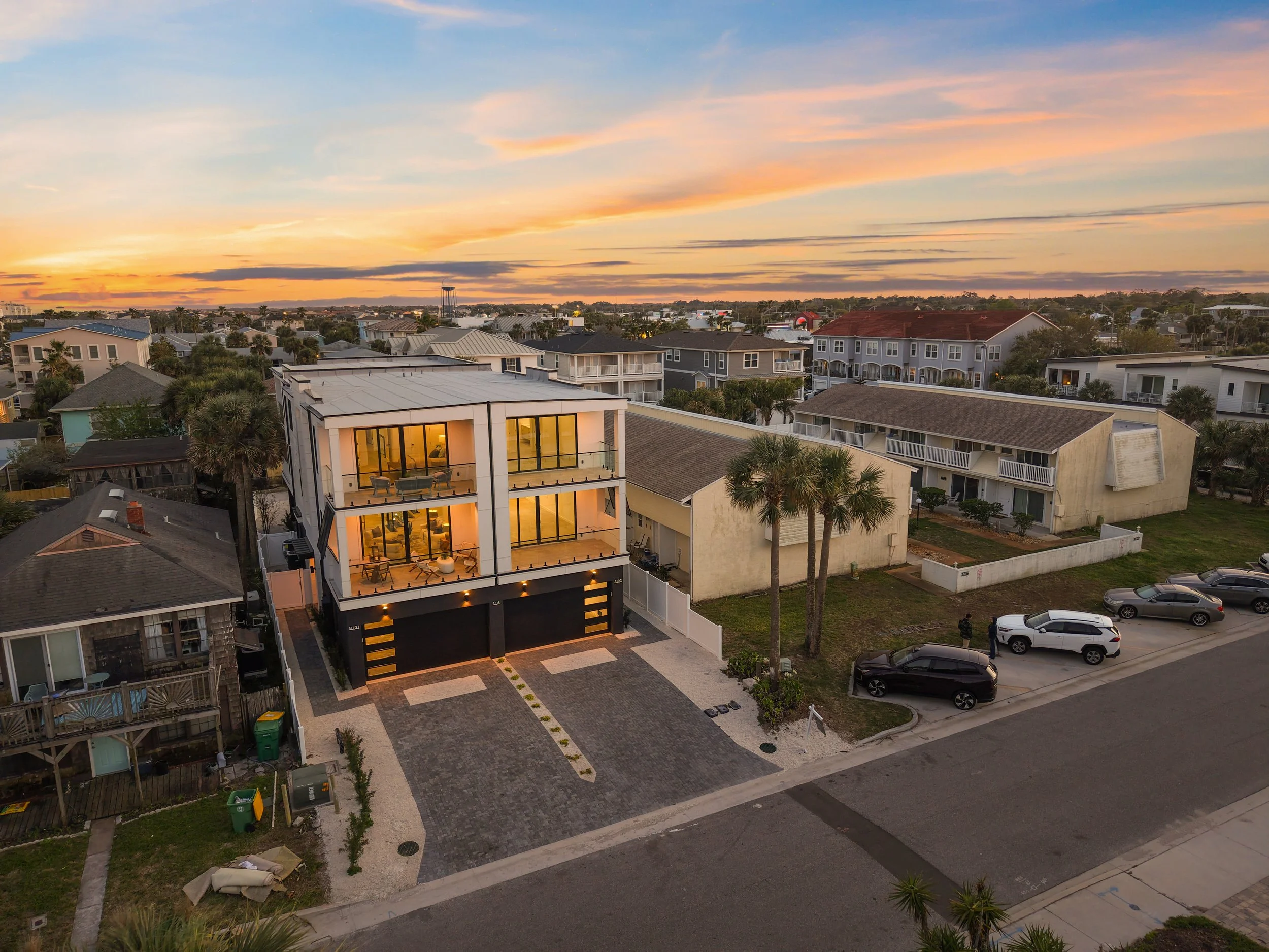 Aerial view of a residential neighborhood during sunset, featuring multiple houses, trees, and parked cars.