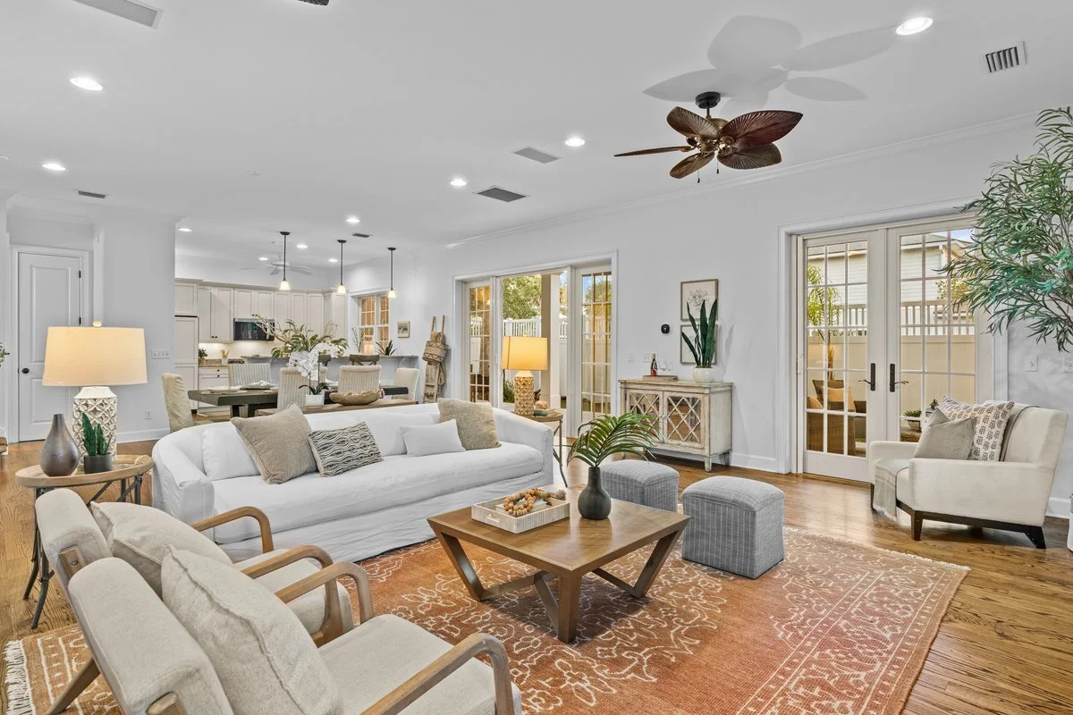 Bright and airy living room with white walls, hardwood floors, a white sofa, armchairs, a wooden coffee table with a tray and plant, side table with table lamp, large windows and glass doors, ceiling fan, and open kitchen visible in the background.