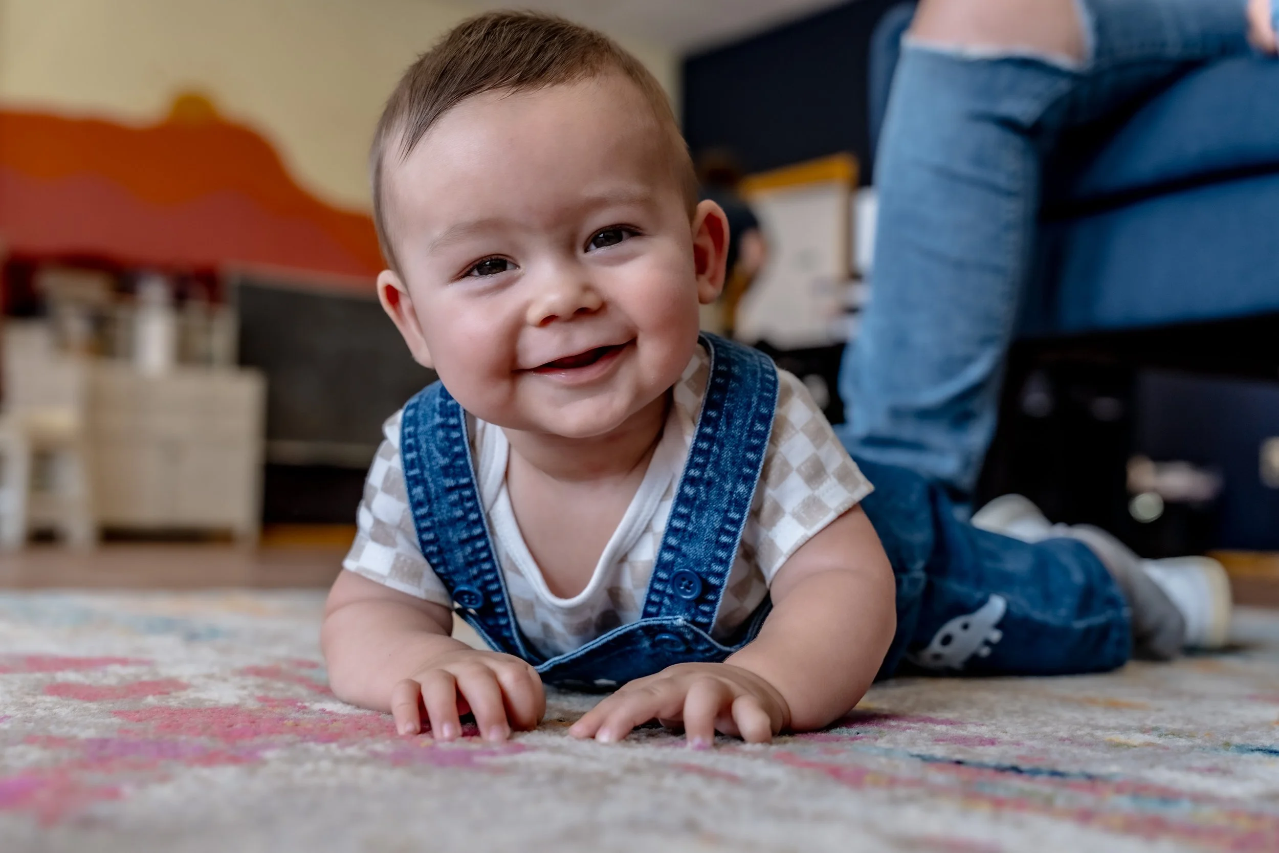 Close-up of a smiling baby with brown hair, lying on a colorful rug, wearing a beige and white checkered shirt and blue denim overalls.