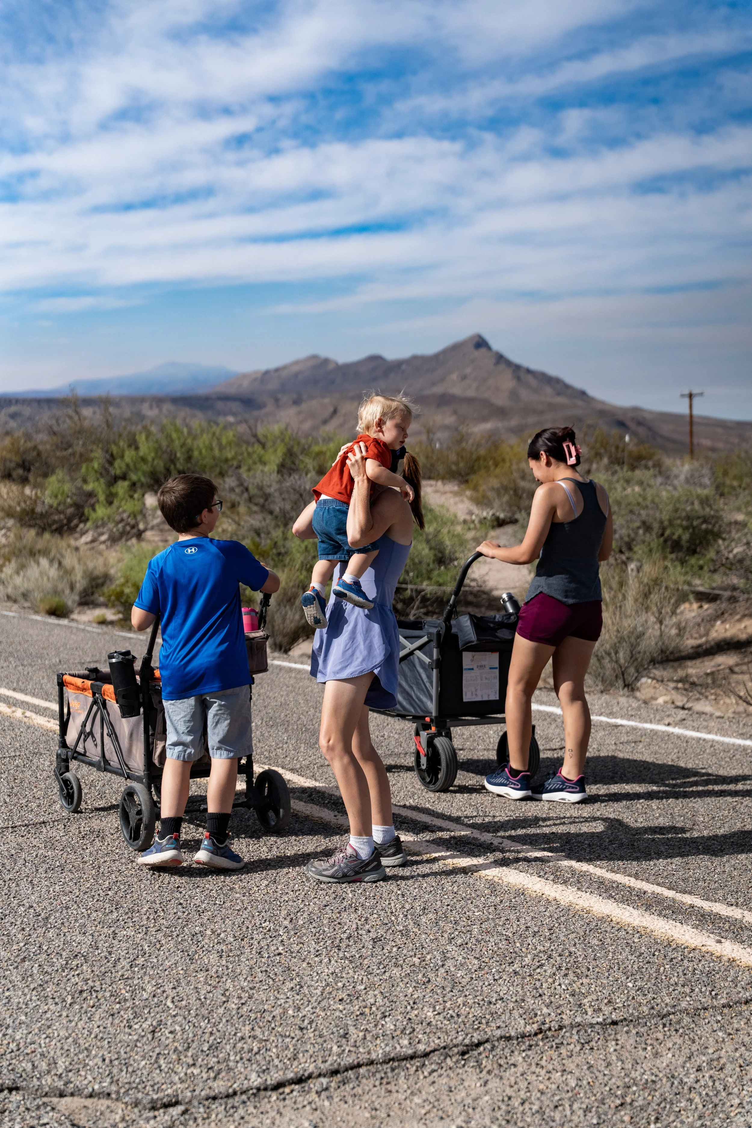 A family of five standing on a rural road against a backdrop of mountains and cloudy sky. One adult carries a young child, another adult pushes a stroller, and two children are with them, one in a blue shirt and the other in a black tank top.